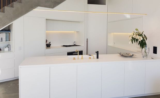 Bright minimalist kitchen with a large white island, integrated cabinetry, concealed under-cabinet lighting, pale stone or concrete finishes, brass tapware, and a black cooktop and sink fixtures. A concrete stairway and open shelving are visible at the left, with a small dining or living area suggested beyond the island.