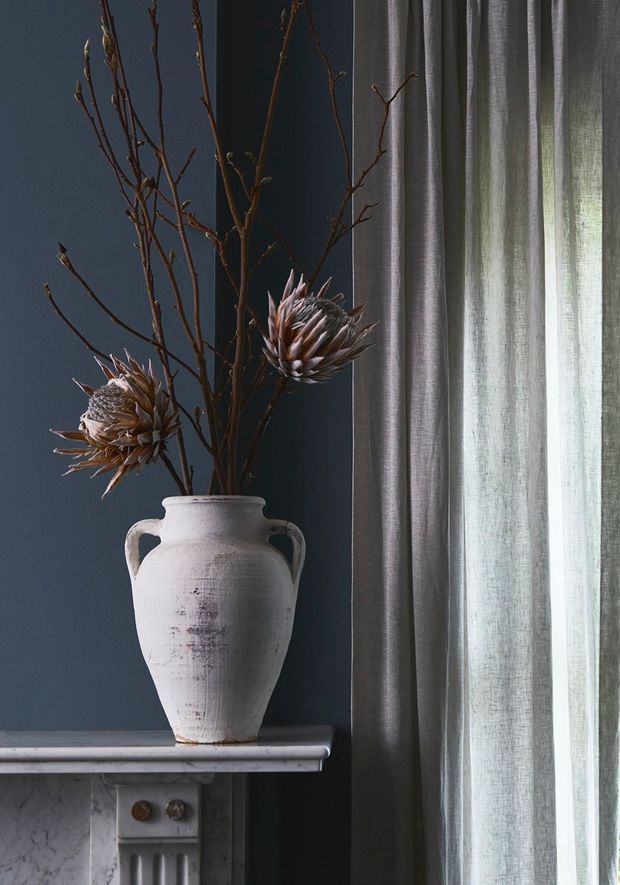 A close-up interior vignette showing a distressed white ceramic urn with dried botanical branches and seed heads on a marble mantel. The backdrop is a deep blue-grey wall beside layered grey sheer and blackout curtains, with soft natural light filtering through the window. Decorative architectural details of the mantel and wall panelling are visible.