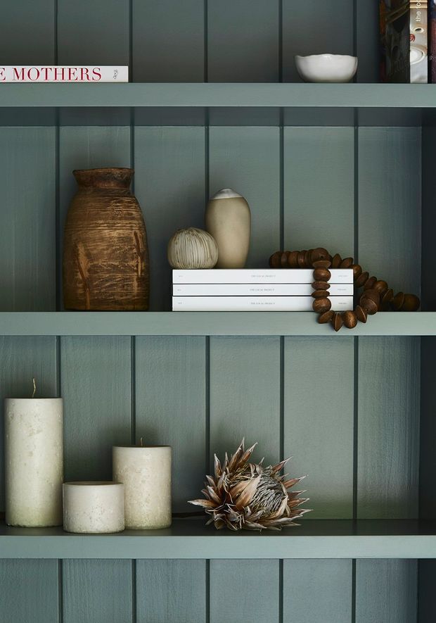 Close-up styled built-in shelving with dark muted green vertical timber wall panelling and matching horizontal shelves. The shelves display books, ceramic vessels, a large rustic earthenware vase, wooden bead decor, candles, and a dried botanical arrangement. The composition is moody, refined, and textural, with no wider room context visible.