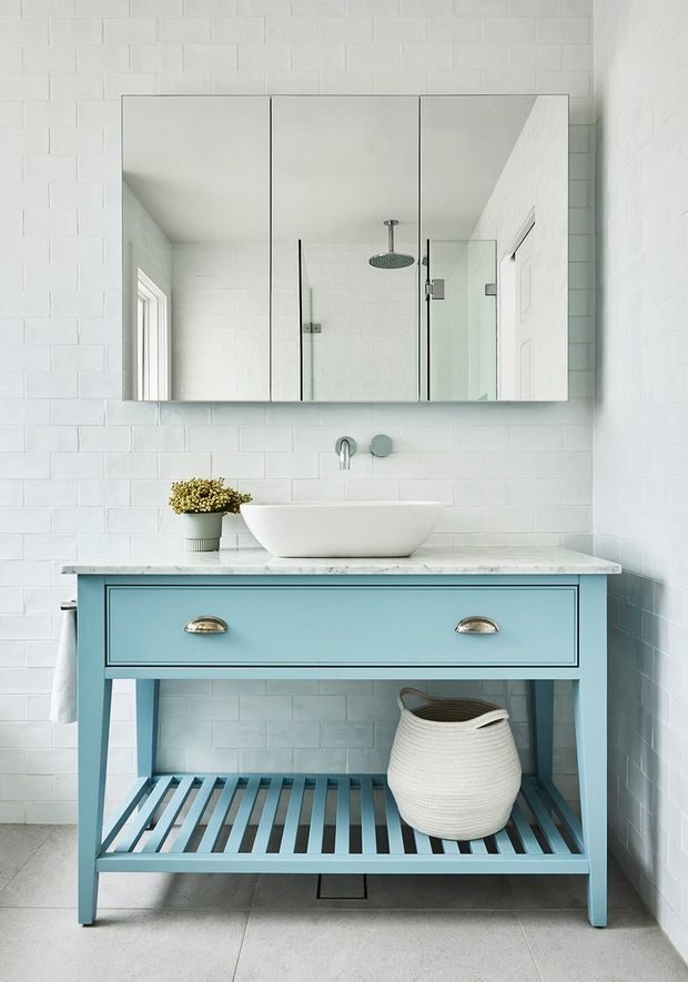 Bright contemporary bathroom featuring white painted brick walls, a large mirrored cabinet, glass-enclosed shower, white vessel basin, and a freestanding light-blue timber vanity with open slatted shelving. The room has a fresh, coastal-inspired palette with marble-look countertop, chrome tapware, and pale tiled flooring.