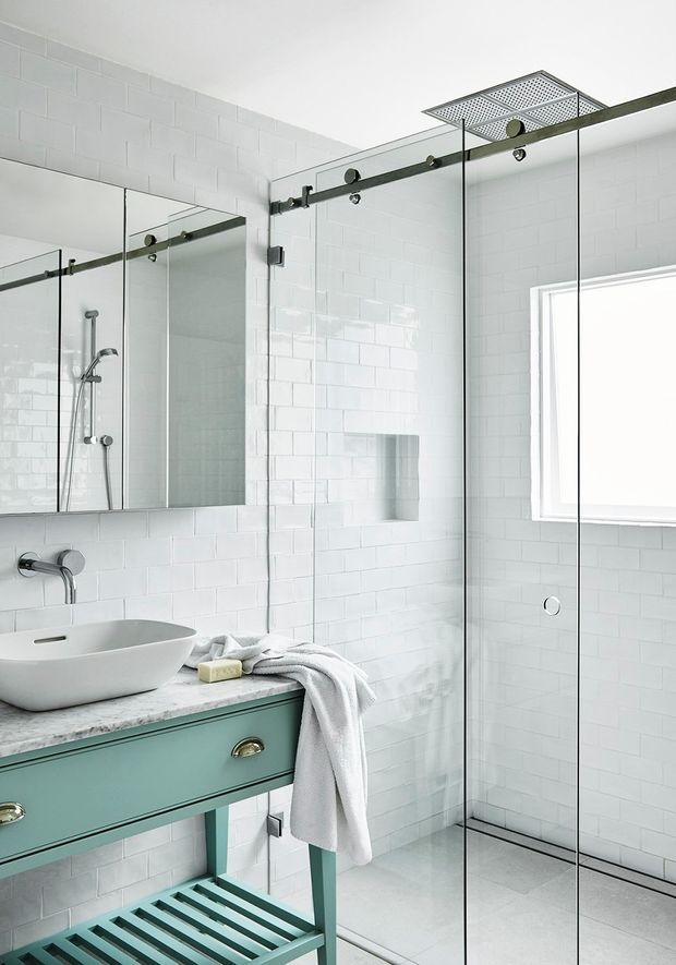 Bright contemporary bathroom featuring white subway-tiled walls, a frameless glass walk-in shower with exposed dark metal sliding hardware and a square rainfall showerhead, a white vessel basin on a mint-green vanity, and a pale stone or marble-look countertop. A large mirror, recessed shower niche, and window enhance the clean, airy space.