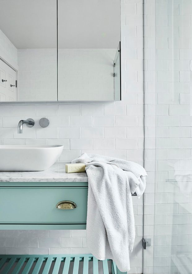 Bright, refined bathroom vanity area featuring white subway tile walls, a frameless glass shower screen, a wall-mounted white vessel basin, pale marble benchtop, and a muted aqua-green vanity drawer with brass cup pull. A white towel and soap rest on the counter, with a large mirror above.