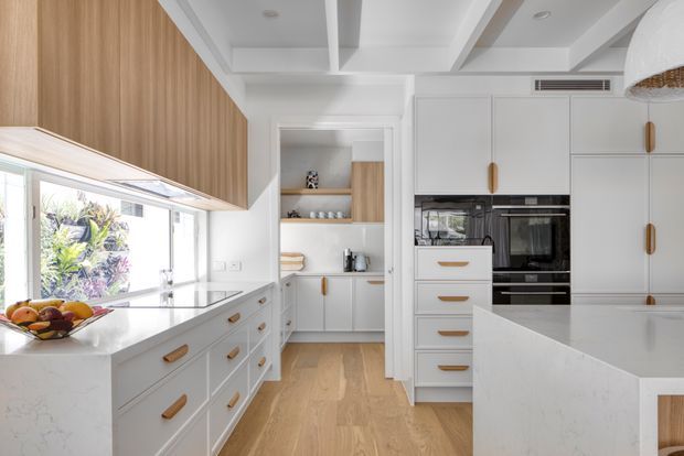 Bright, refined contemporary kitchen with white Shaker-style cabinetry, warm timber veneer overhead joinery, pale stone benchtops, integrated black appliances, and timber flooring. The room features a large island, extensive storage, a window overlooking greenery, and a coffered ceiling with recessed lighting and a woven pendant.