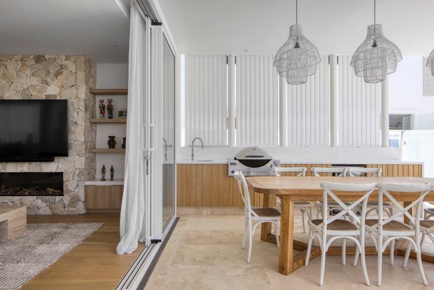 Bright open-plan dining and kitchen interior with a living area visible to the left through a wide glazed opening. The room features a large timber dining table with white cross-back chairs, a timber-fronted kitchen island or cabinetry, pale stone-look flooring, white vertical blinds, woven pendant lights, and a prominent rough natural-stone fireplace wall with television and built-in shelving.