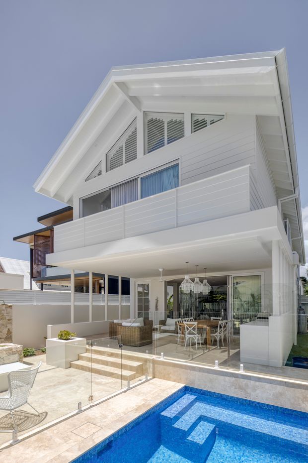 Bright exterior view of a multi-level white coastal-style residence with a steep gabled roof, horizontal cladding, louvred upper windows, and a covered outdoor entertaining area opening onto a tiled terrace and vivid blue swimming pool. Glass pool fencing, outdoor dining furniture, and a partially enclosed balcony are visible.