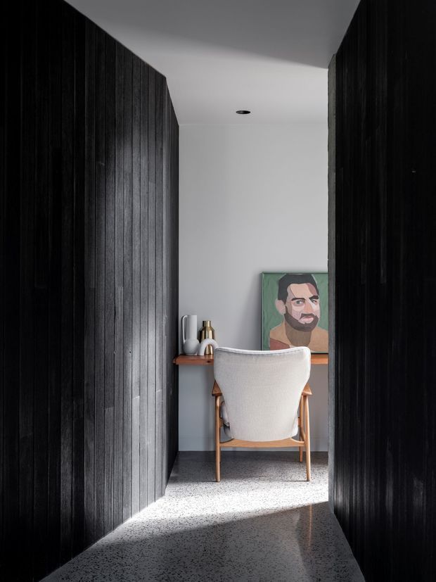 A narrow, highly contrasted home office or study nook viewed from a dark corridor. Black vertical timber wall panelling lines both sides, opening onto a white-plastered workspace with a simple wooden desk, upholstered desk chair, decorative objects and a colourful portrait artwork. A polished speckled terrazzo floor extends through the space, with daylight creating a bright diagonal patch across the floor.