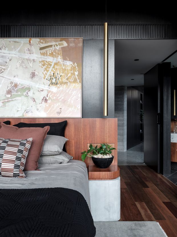 A moody contemporary bedroom with a low timber bedhead and integrated side ledge, layered grey bedding, black throw, and muted burgundy and patterned cushions. A large abstract artwork hangs above the bed against a dark vertically ribbed wall, complemented by a slim brass pendant light. Dark timber flooring leads toward a shadowy hallway and adjacent built-in cabinetry, while a concrete-toned bedside block and potted plant add texture.