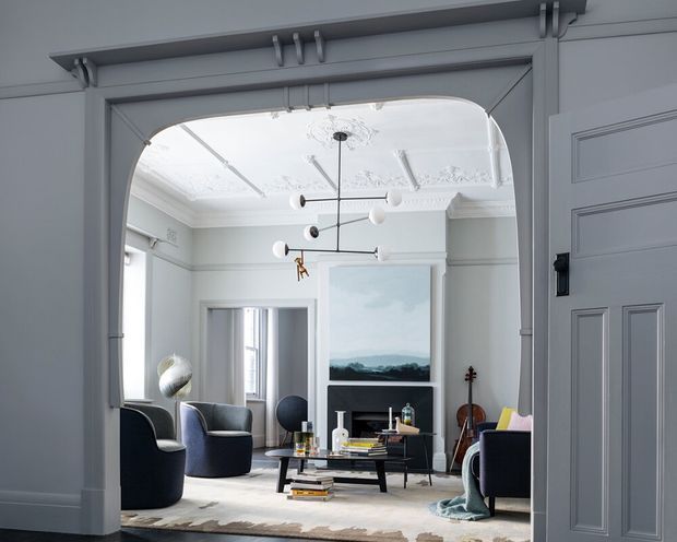 A refined living room viewed through a wide arched opening, featuring pale grey-blue walls and trim, ornate decorative ceiling plasterwork, a contemporary sculptural pendant light, dark upholstered lounge chairs, a low black coffee table, a fireplace or media wall, artwork, and a violin. The space combines classical architectural detailing with contemporary furnishings.
