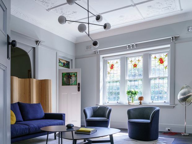 Refined living room with pale grey walls, ornate plaster ceiling panels and cornices, stained-glass windows and door inserts, and dark blue upholstered seating arranged around layered oval coffee tables. A sculptural modern pendant contrasts with the period detailing, while a curved timber screen and decorative floor lamp add visual interest.