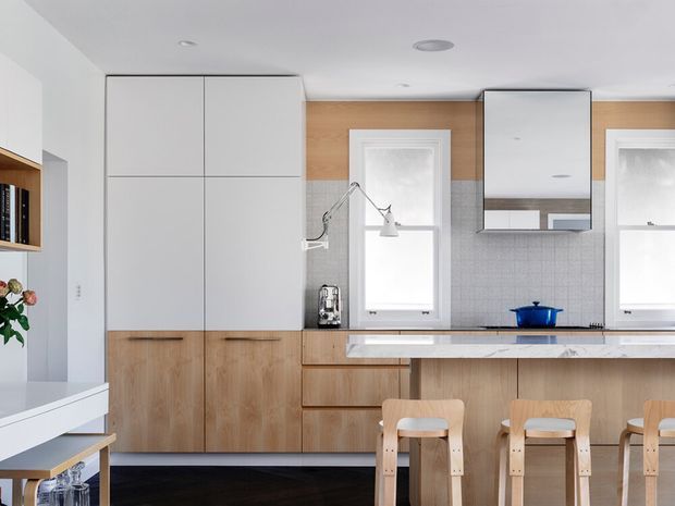 Bright contemporary kitchen with extensive light timber cabinetry, a pale stone benchtop, and a tiled backsplash. The room features a central island with integrated seating, white upper cabinetry, a reflective rangehood, and two tall windows. A glimpse of adjacent dining or living space is visible at left.