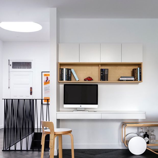 Bright minimalist living and media area with a wall-mounted white built-in entertainment unit, timber open bookshelf, computer monitor, and long floating console. A small timber chair and table sit beside a black metal stair or entry balustrade, with a white entry door and framed artwork. The room has white walls and cabinetry, pale timber joinery, and dark herringbone timber flooring.