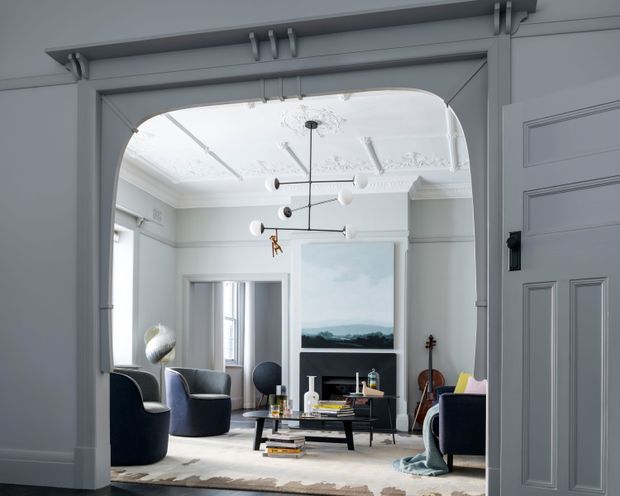 A refined living room viewed through a large arched opening, with pale grey-blue walls, ornate decorative ceiling mouldings, dark upholstered armchairs, a low black coffee table, a fireplace or media wall, contemporary pendant lighting, artwork, and a violin. A paneled door and substantial trim frame the foreground.