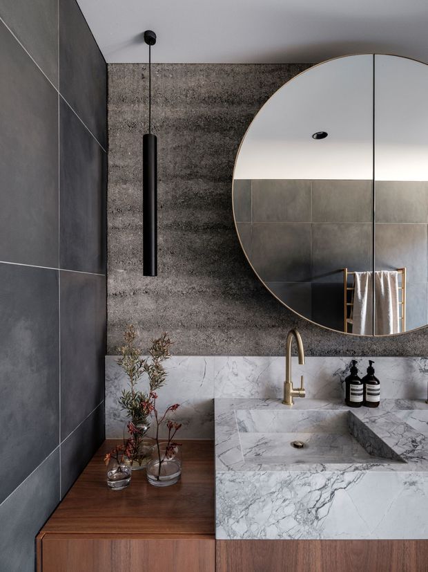 A refined contemporary bathroom vanity with a large round mirror, dark grey wall and floor-to-ceiling tiles, a textured charcoal feature wall, and a white-grey veined stone benchtop with integrated rectangular basin. Warm timber cabinetry and a brushed brass tap add contrast, while a slim black pendant and minimal accessories complete the moody, luxurious scheme.