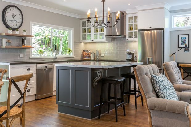 A bright, traditionally detailed open-plan kitchen with a dark painted island, white shaker cabinetry, stainless-steel appliances, pale subway-tile splashback, timber flooring, and a large upholstered seating area. A decorative chandelier, glass-front cabinets, crown moulding, and vintage-style wall clock add a refined, classic character.
