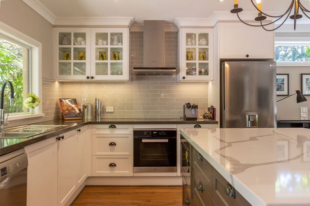 A warm, traditional-style kitchen with cream painted cabinetry, glass-front upper cupboards, a tiled splashback, stainless-steel appliances, dark stone-look benchtops, and a large central island with a pale polished surface. Timber flooring and a decorative chandelier add warmth and character, while a window brings in natural light.