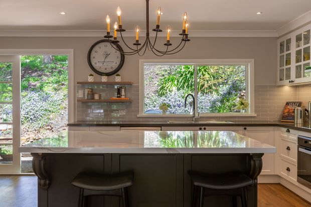 A warm, traditional-style kitchen centred on a large island with a dark fixed base, pale stone benchtop and two stools. The room features shaker-style cabinetry, a glossy rectangular tile splashback, timber flooring, white-framed windows, a built-in oven, open shelving, and a prominent multi-arm chandelier. Lush vegetation is visible outside the windows.