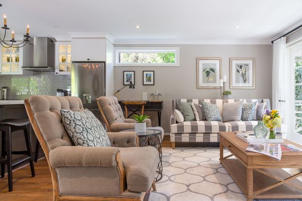 A bright, comfortably furnished open-plan living room with a connected kitchen visible at left. The space features a pair of plush upholstered armchairs, a patterned sofa, timber coffee table, pale geometric rug, framed botanical artwork, pendant and recessed lighting, and large windows and glazed doors. The kitchen includes light cabinetry, a stainless-steel refrigerator, and a green-tile backsplash. The overall look is refined, warm, and transitional with traditional detailing.