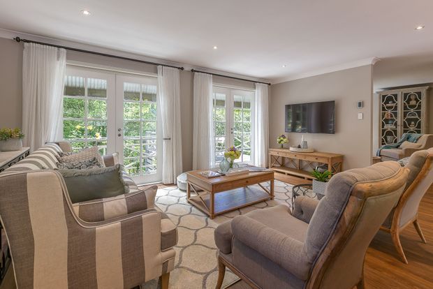 A bright, traditionally styled living room with multiple upholstered armchairs and sofas arranged around a timber coffee table. Full-height French doors with sheer curtains open to greenery, while a wall-mounted television, timber media console, patterned area rug, recessed ceiling lights, and decorative built-in cabinetry complete the room.