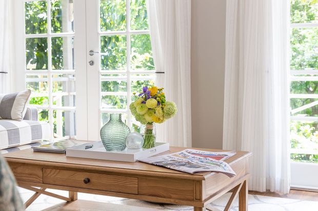 Bright, airy living room vignette featuring a mid-century-inspired wooden console table with drawers and crossed supports, styled with books, magazines, glass decor and a floral arrangement. White multi-pane French doors and large windows with sheer curtains create strong natural light; a sofa is partially visible at left.