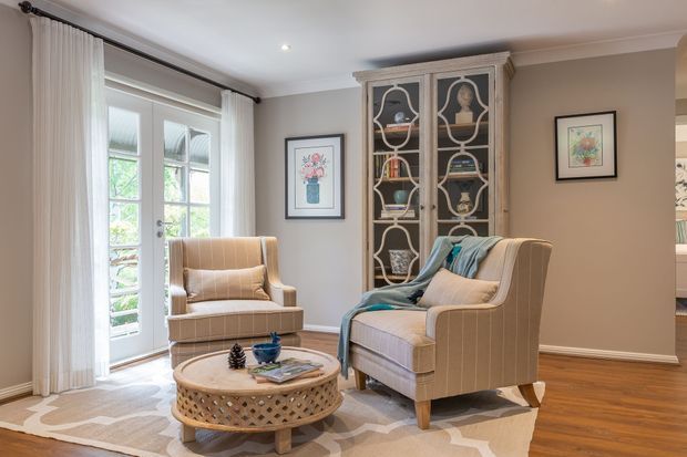 A refined sitting area with two upholstered armchairs arranged around a round woven coffee table on a patterned rug. French doors with sheer curtains open to greenery, while a tall glass-front display cabinet, framed artwork, warm neutral walls, white trim, and timber flooring create a traditional, inviting interior.