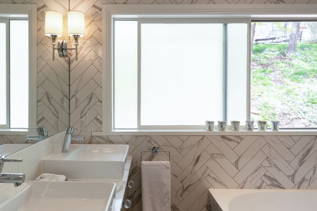 Bright, refined bathroom with a double vanity, large white vessel-style basins, chrome fittings, a freestanding bath partially visible at right, and a large window overlooking greenery. The walls feature a striking pale marble-look herringbone tile pattern, complemented by a decorative wall sconce and small potted plants along the window sill.