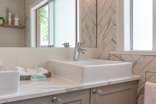 Bright, refined bathroom vanity featuring a white vessel sink on a pale stone benchtop, grey painted cabinetry with crystal-style knobs, chrome tapware, and a geometric herringbone-pattern tiled wall. Natural light enters through the window, with neatly arranged towels and toiletries adding a soft, welcoming touch.
