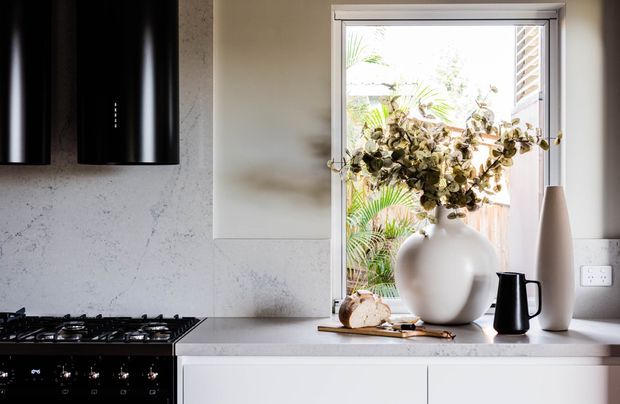 Bright contemporary kitchen vignette with a black gas cooktop and oven, white engineered-stone-style splashback and benchtop, pale cabinetry, matte black pendant lights, and a window styled with a large white vase, dried foliage, and black vessels.