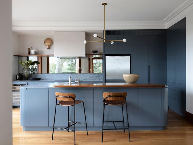 A refined contemporary kitchen with a large blue-grey island, timber benchtop, integrated stainless-steel appliances, dark blue full-height cabinetry, and two timber-and-black bar stools. Warm timber flooring, white decorative ceiling mouldings, brass pendant lighting, and a window overlooking greenery create a balanced, sophisticated interior.