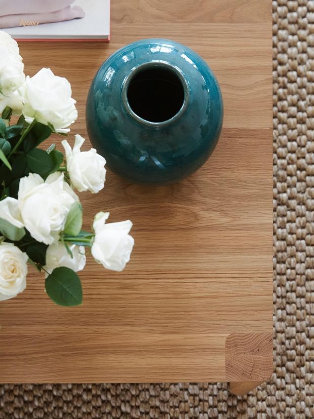 Top-down still life of a warm natural timber coffee table with visible grain and rounded corner, placed on a textured woven rug. A glossy deep teal ceramic vase and white flowers sit on the table beside a partially visible book, creating a refined, organic interior styling vignette.