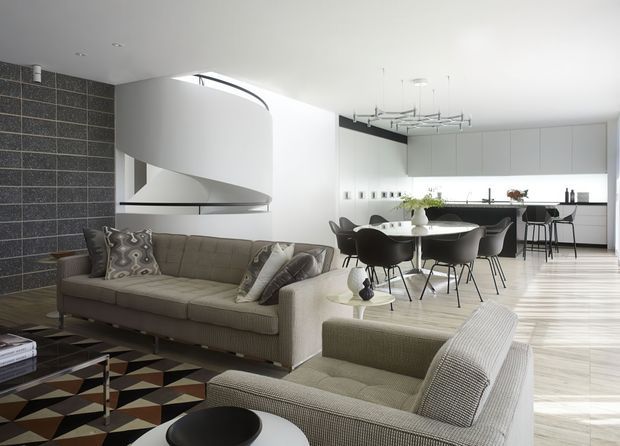 A bright, open-plan contemporary interior combining a living room, dining area, and kitchen. The room features two neutral upholstered sofas, a geometric patterned rug, a dark tiled feature wall, a sculptural curved white fireplace or architectural volume, a glass dining table with dark chairs, minimalist white kitchen cabinetry, and a linear sculptural pendant light.