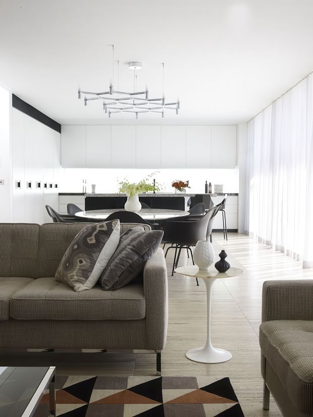 Bright open-plan contemporary interior featuring a living room in the foreground and a dining area with a minimalist white kitchen behind it. The space has pale wood flooring, extensive white cabinetry, sheer full-height curtains, a sculptural linear pendant light, dark dining chairs, a textured neutral sofa, and a geometric patterned rug.