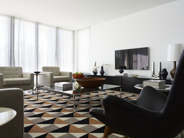 Bright, refined living room with floor-to-ceiling sheer curtains, pale upholstered lounge chairs, a black leather sculptural armchair, glass coffee table, dark media console, decorative ceramics, and a bold geometric patterned rug. The space has a clean contemporary shell with restrained black-and-white furnishings and warm earthy rug accents.