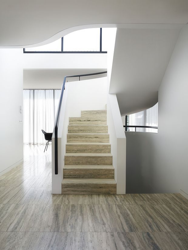 Bright contemporary stairway with broad pale stone or timber-look tiled steps, a slim black metal handrail, white walls, and sculptural curved ceiling and stair forms. Floor-to-ceiling glazing and clerestory windows bring in abundant natural light, with a glimpse of an adjacent open living area and sheer curtains.