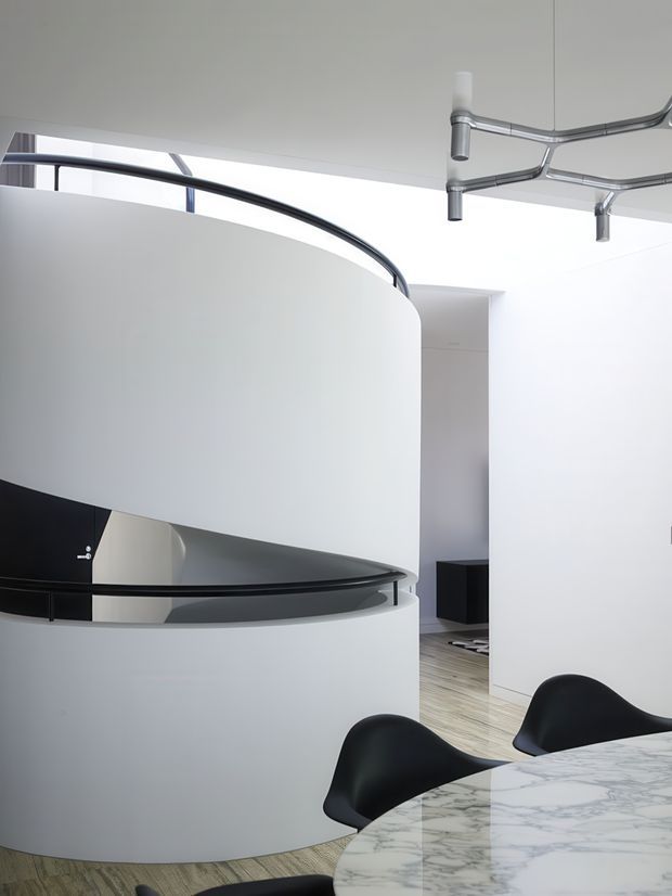 A bright, minimalist contemporary interior featuring a sculptural curved white staircase or mezzanine enclosure with sleek black railings. A polished marble dining table with black molded chairs occupies the foreground, while a modern exposed metal ceiling track or services installation adds an industrial accent. Pale timber flooring, white walls, and strong curved forms create an open, refined space.
