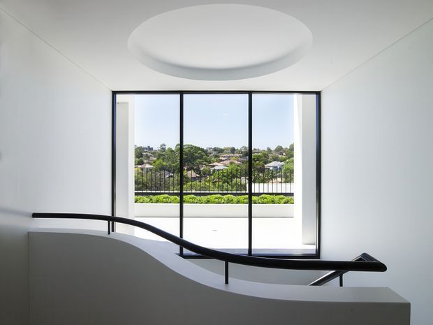 A minimalist stairway or landing with a sculptural white curved balustrade and black metal handrail. Full-height black-framed glazing opens to a balcony or terrace with a suburban view beyond. The space has crisp white walls and ceiling, a large circular flush-mounted ceiling light, and strong natural daylight.