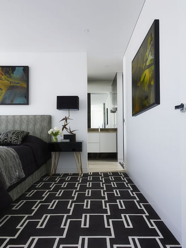A narrow, contemporary interior corridor or living area with a dark patterned carpet, white walls and ceiling, and minimalist black furnishings. A grey upholstered sofa with black throws sits along the left wall beside a slim console table, black lamp, decorative object, and flowers. Framed artwork hangs on both walls, while a compact white kitchenette or utility area is visible at the rear through the passage.