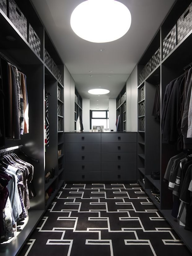 A dark, highly organised walk-in wardrobe with floor-to-ceiling open shelving and hanging rails on both sides, central matte dark drawer cabinetry, patterned storage baskets, and a bold black-and-white geometric floor covering. Circular recessed ceiling lights provide bright, focused illumination, while a mirror and small window are visible at the far end.