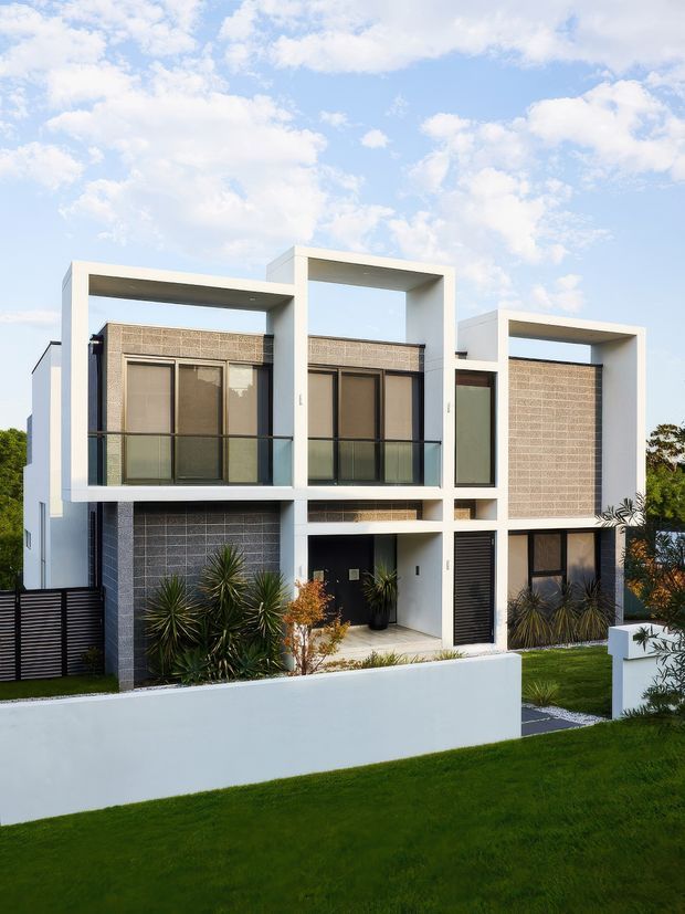 Contemporary multi-storey residential facade with crisp white geometric frames, dark window and door surrounds, textured grey masonry panels, projecting balconies with glass balustrades, and a landscaped front garden behind a low white boundary wall.