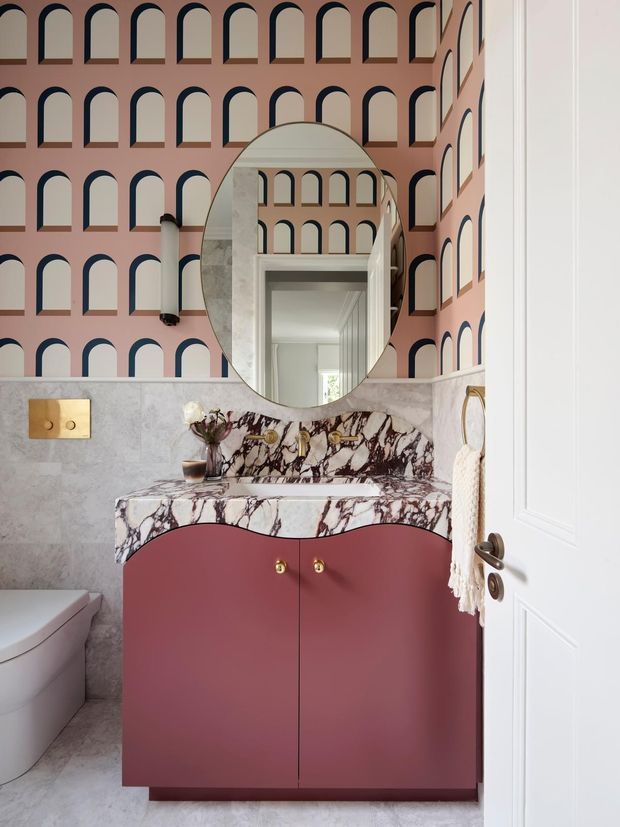 A compact, highly styled bathroom featuring a burgundy curved-front vanity with gold knobs, dramatic veined marble benchtop and splashback, pale grey stone-look wall and floor finishes, and a large oval mirror. Pink wallpaper with repeating navy-trimmed arched motifs creates a bold feature wall, while a white toilet and paneled door are visible at the edges.