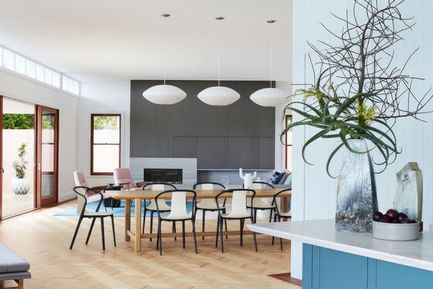 Bright open-plan kitchen and dining area with a timber herringbone floor, light walls, dark built-in kitchen cabinetry, pale benchtops, and a large dining table surrounded by contemporary chairs. Floor-to-ceiling glazing and timber-framed doors bring in natural light, while sculptural pendant lights and a large decorative vase add visual interest. A blue kitchen island or joinery element is visible in the foreground.