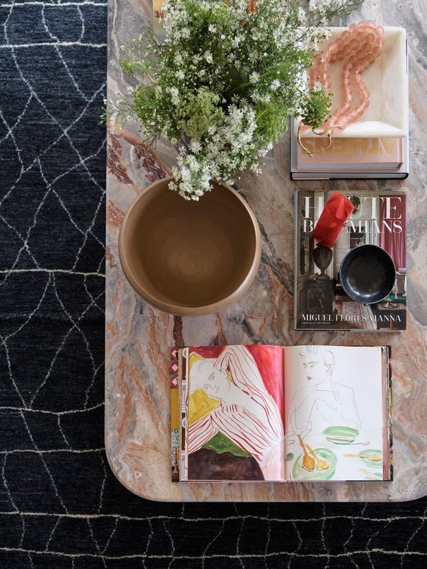 Top-down view of a styled coffee table with a richly veined stone surface and rounded corners, placed over a dark patterned rug. The table holds a large ceramic bowl, flowers, stacked art and design books, a small dark bowl, decorative beads, and an open sketchbook. The composition has a curated, artistic interior styling aesthetic.
