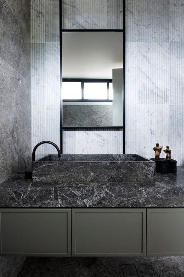 A moody, refined bathroom vanity featuring a dark veined natural-stone benchtop and integrated raised basin, matte dark cabinetry, black tapware, and a tall black-framed mirror. Grey stone wall cladding and flooring create a dramatic, monochromatic material palette, with small sculptural objects placed beside the basin.