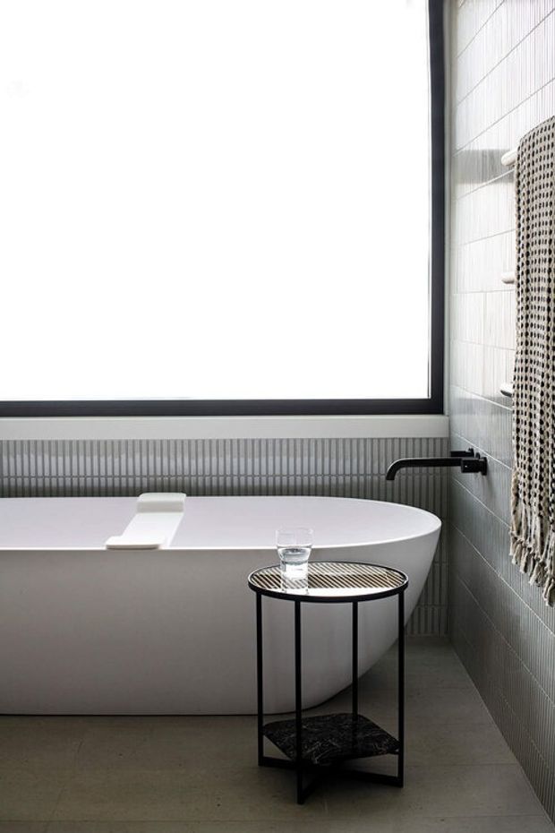 A refined contemporary bathroom featuring a freestanding white oval bathtub beneath a large black-framed window. The walls are clad in small light grey tiles with a vertically ribbed tile detail behind the bath, complemented by a matte dark floor. A minimalist black wall-mounted tap and a slim round black metal side table complete the calm, monochromatic scheme. A textured hanging textile is visible at the right edge.