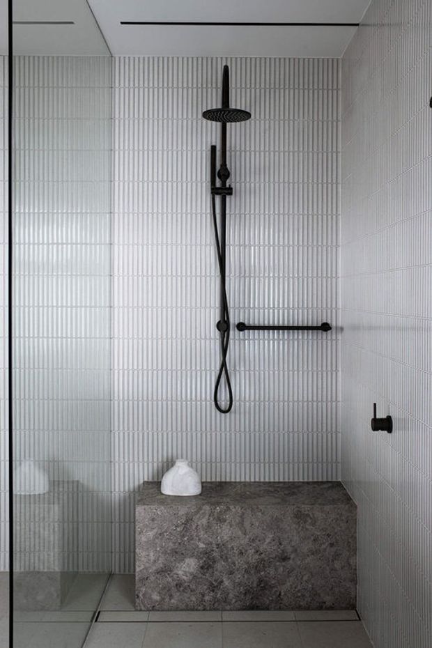 Minimalist walk-in shower with full-height white vertically fluted tile walls, black rain shower and handheld fittings, clear glass screen, and a substantial grey stone-look built-in bench. The palette is monochromatic with strong black hardware accents and clean contemporary detailing.
