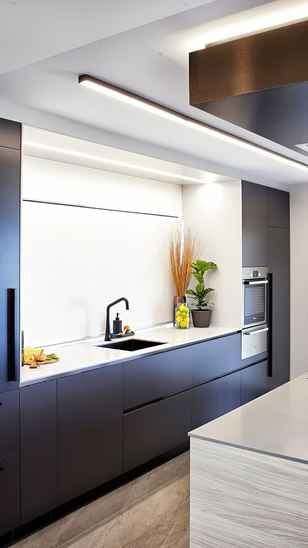 A sleek contemporary kitchen with full-height dark cabinetry, white integrated wall cabinetry and benchtops, a black undermount sink with sculptural tap, and a built-in oven. Linear LED lighting runs through the white ceiling, while a dark bronze-toned ceiling feature adds contrast. A pale wood-grain island occupies the foreground, and potted greenery and dried grasses add natural accents.
