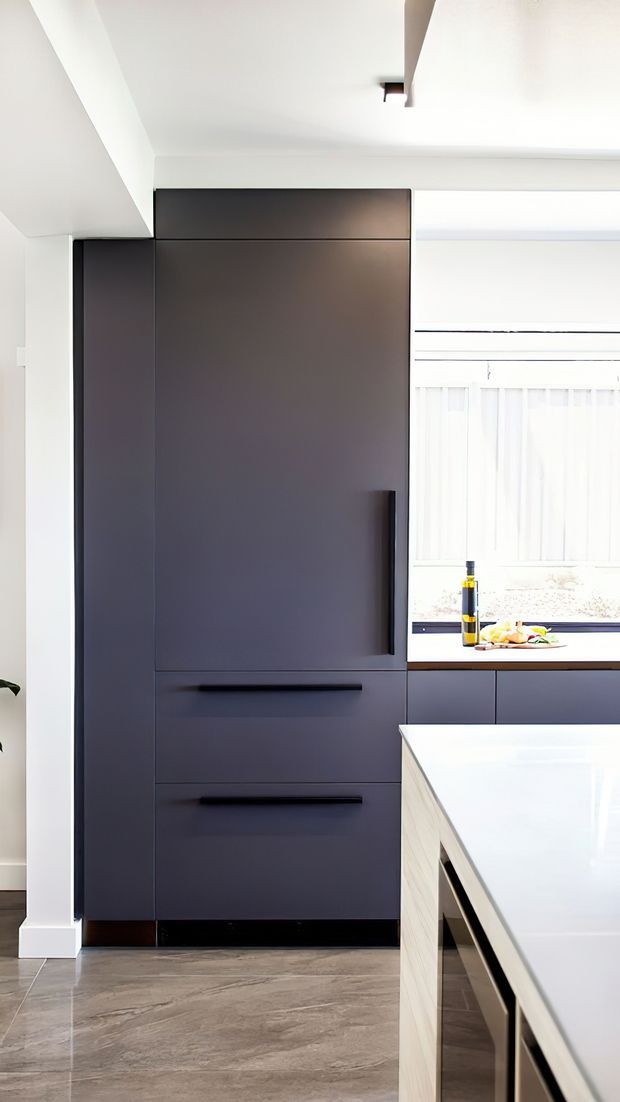 A bright contemporary kitchen interior featuring a tall bank of matte dark cabinetry with integrated handles and drawers, a large pale island with a light benchtop, a window with sheer blinds, and large-format grey stone-look flooring. The space has clean lines, restrained detailing, and strong natural light.