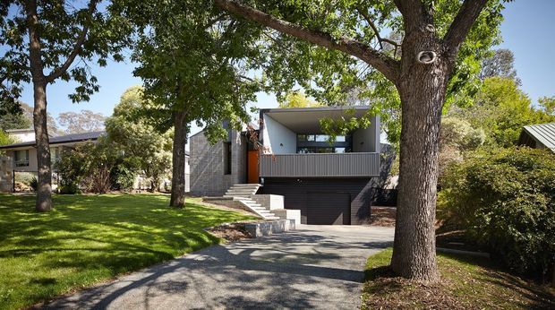 Exterior view of a contemporary multi-level home set among mature trees and landscaped lawn. The house features a strong geometric form, a flat roof, dark horizontal garage door, concrete stairway, grey masonry walls, and a covered upper-level balcony or terrace with metal balustrading. A paved driveway leads toward the integrated garage.
