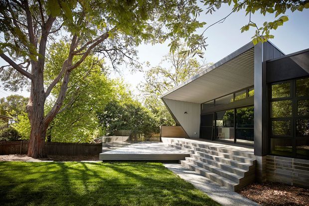 Exterior view of a contemporary single-storey home set within a leafy garden. The house features dark framed glazing, a dramatic projecting skillion roof with a soffit, grey masonry cladding, broad steps, and a raised timber deck or terrace overlooking the lawn.