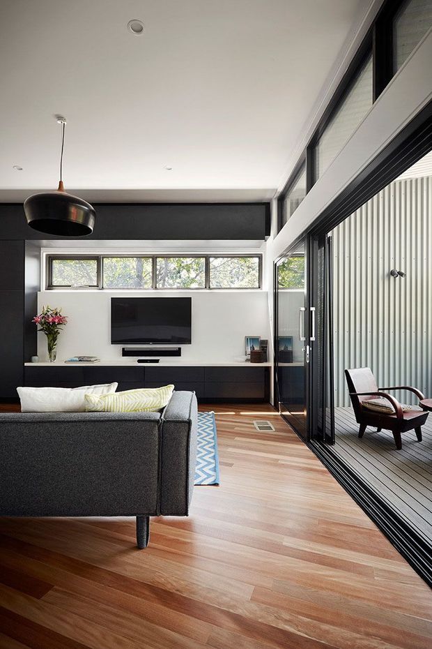 A contemporary living room with a dark grey sofa facing a wall-mounted television and low black media joinery. Warm timber flooring contrasts with black and white built-in surfaces, clerestory windows, and extensive black-framed glazing opening to a covered outdoor deck. A sculptural pendant light, patterned rug, and timber-framed lounge chairs add detail.