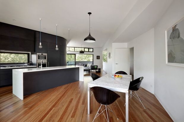 Open-plan contemporary kitchen and dining area with dark integrated cabinetry, a large white-edged island, timber flooring, marble-look dining table, black pendant lights, and black dining chairs. Living space and extensive glazing are visible beyond the kitchen.